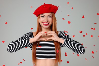 Young woman making heart with hands under confetti on light grey background