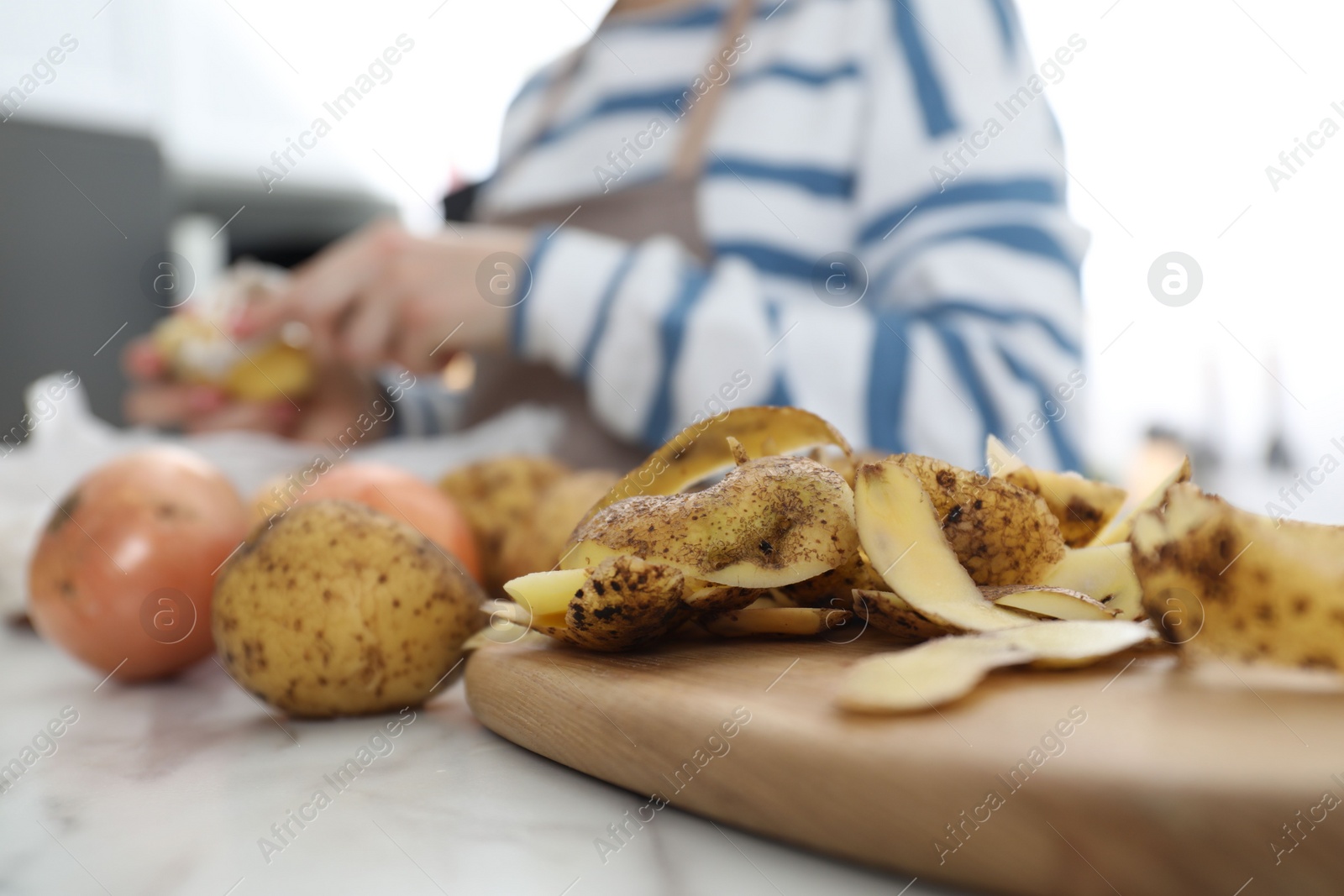 Photo of Woman peeling fresh potato at white marble table indoors, focus on peels