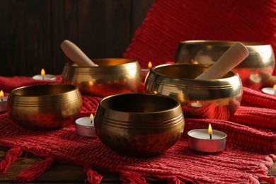 Photo of Tibetan singing bowls with mallets, burning candles and red fabric on wooden table