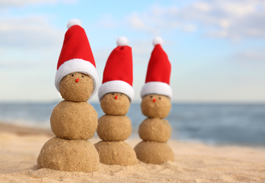Photo of Snowmen made of sand with Santa hats on beach near sea. Christmas vacation
