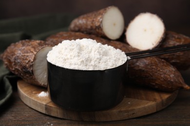 Scoop with cassava flour and roots on wooden table, closeup