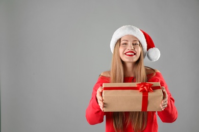 Young beautiful woman in Santa hat with gift box on grey background. Christmas celebration