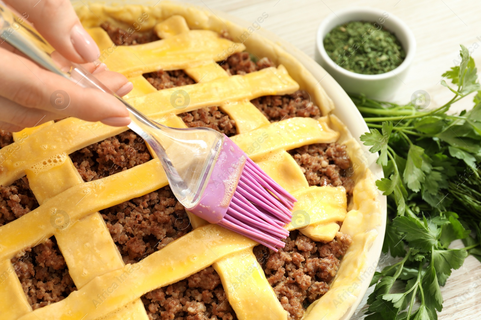 Photo of Woman spreading egg yolk onto raw meat pie at white wooden table, closeup