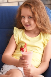 Little girl with glass of delicious milk shake in armchair indoors