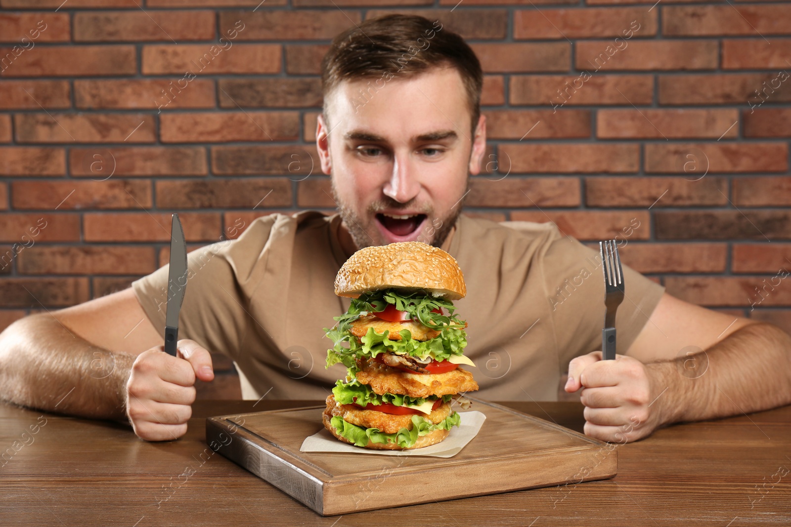 Photo of Young hungry man with cutlery eating huge burger at table