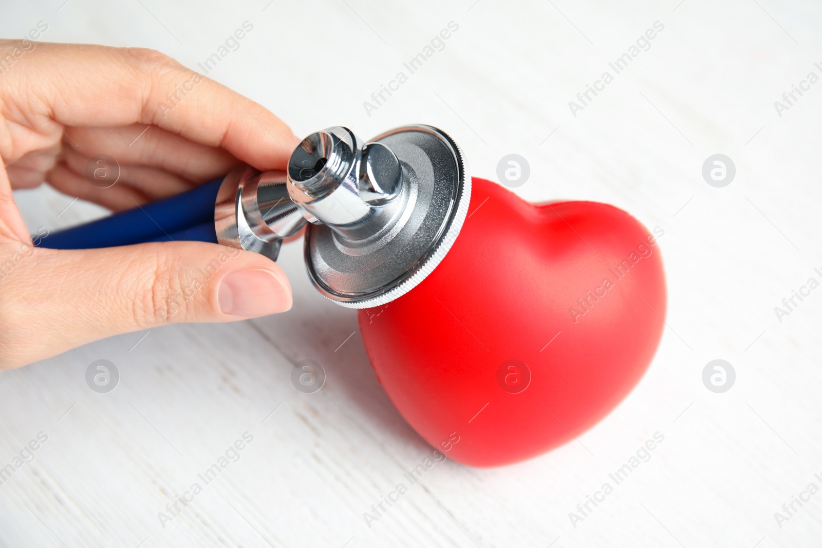 Photo of Woman holding stethoscope near red heart on wooden table, closeup. Cardiology concept