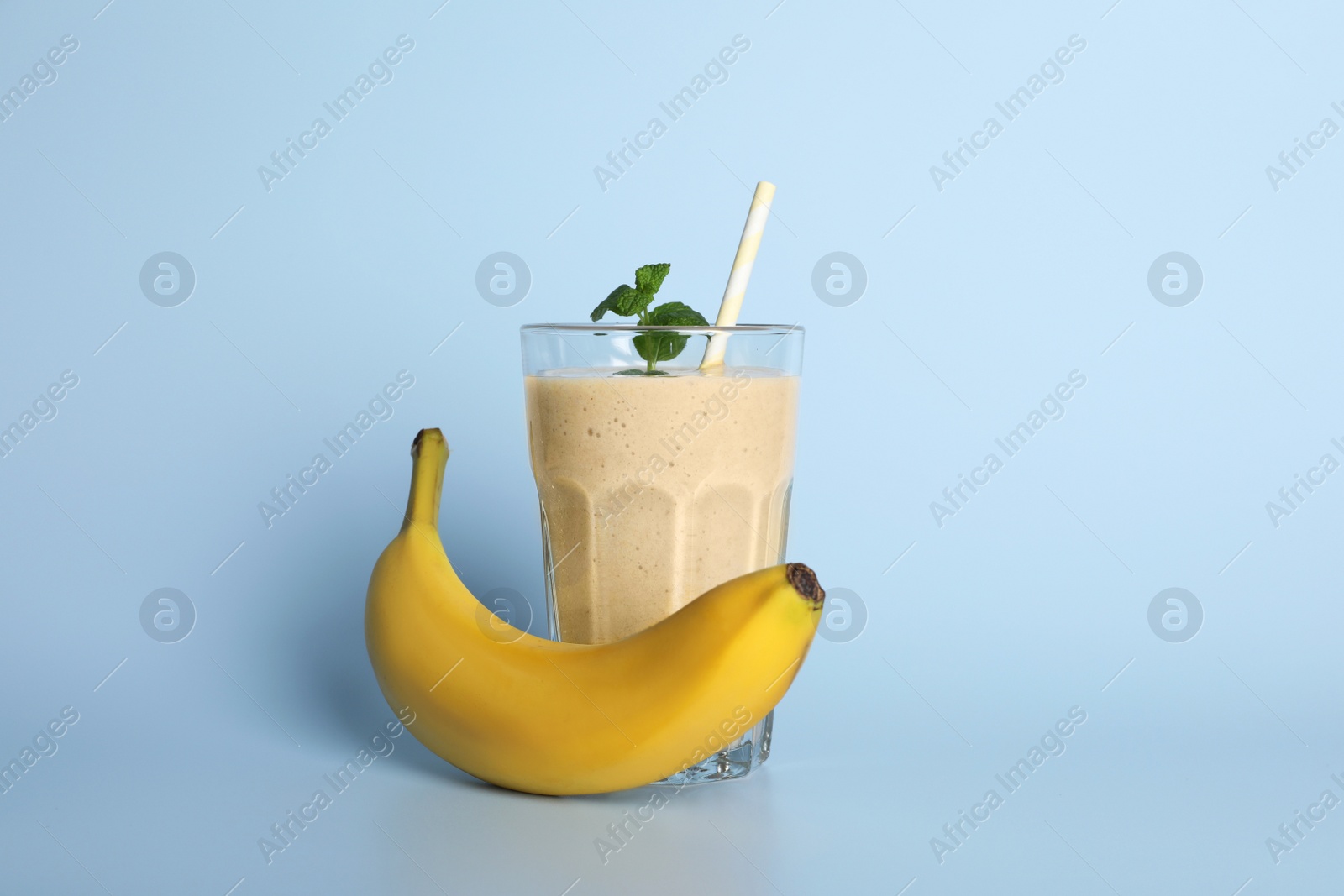 Photo of Glass of tasty smoothie with straw, banana and mint leaves on light blue background