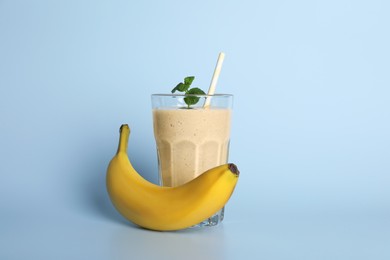 Photo of Glass of tasty smoothie with straw, banana and mint leaves on light blue background