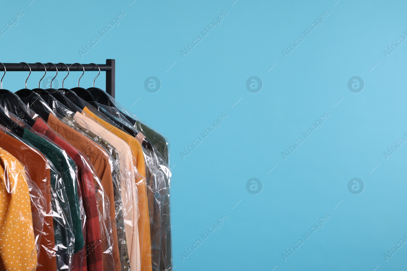 Photo of Dry-cleaning service. Many different clothes in plastic bags hanging on rack against light blue background, space for text