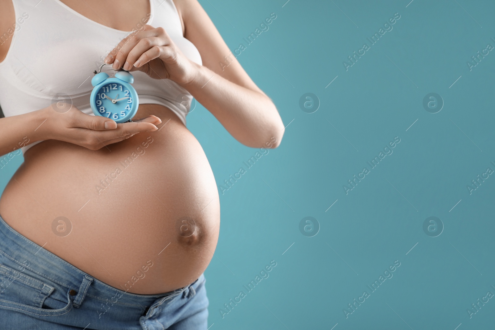 Photo of Young pregnant woman holding alarm clock near her belly on light blue background, closeup and space for text. Time to give birth