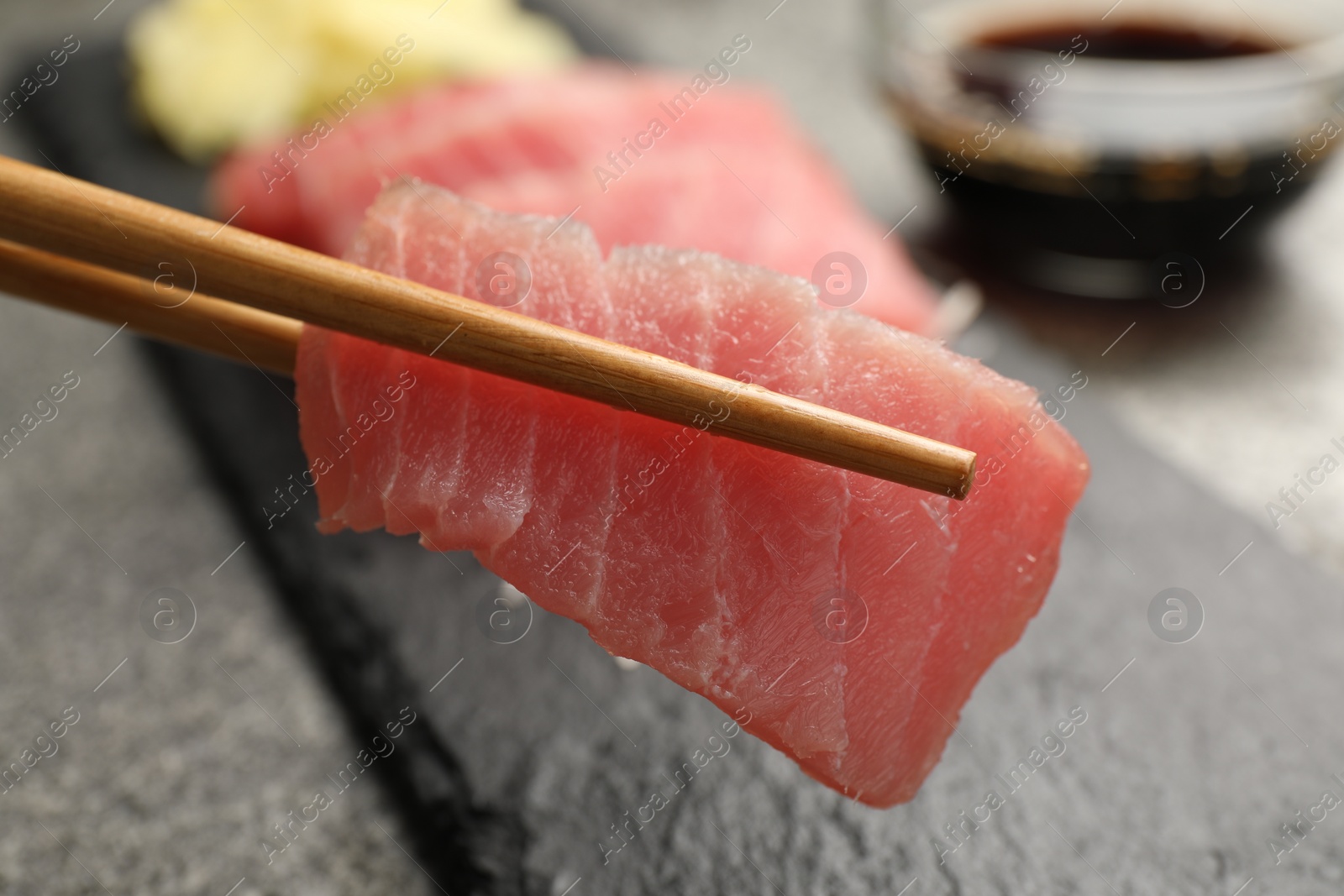 Photo of Holding tasty sashimi (piece of fresh raw tuna) with chopsticks against blurred background, closeup