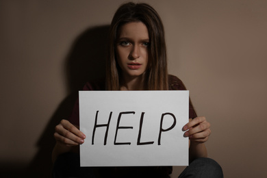 Abused young woman with sign HELP near beige wall. Domestic violence concept