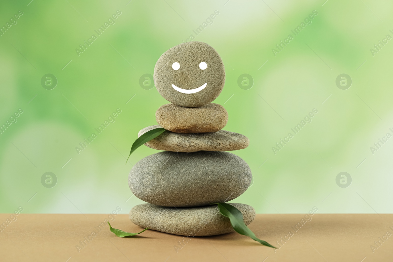 Photo of Stack of stones with drawn happy face and green leaves on table against blurred background. Zen concept