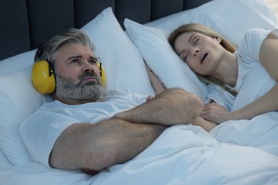 Photo of Irritated man with headphones lying near his snoring wife in bed at home