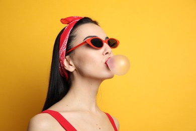 Photo of Fashionable young woman in pin up outfit blowing bubblegum on yellow background, space for text