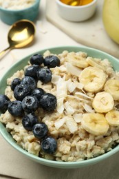 Photo of Tasty oatmeal with banana, blueberries, coconut flakes and honey served in bowl on beige table, closeup