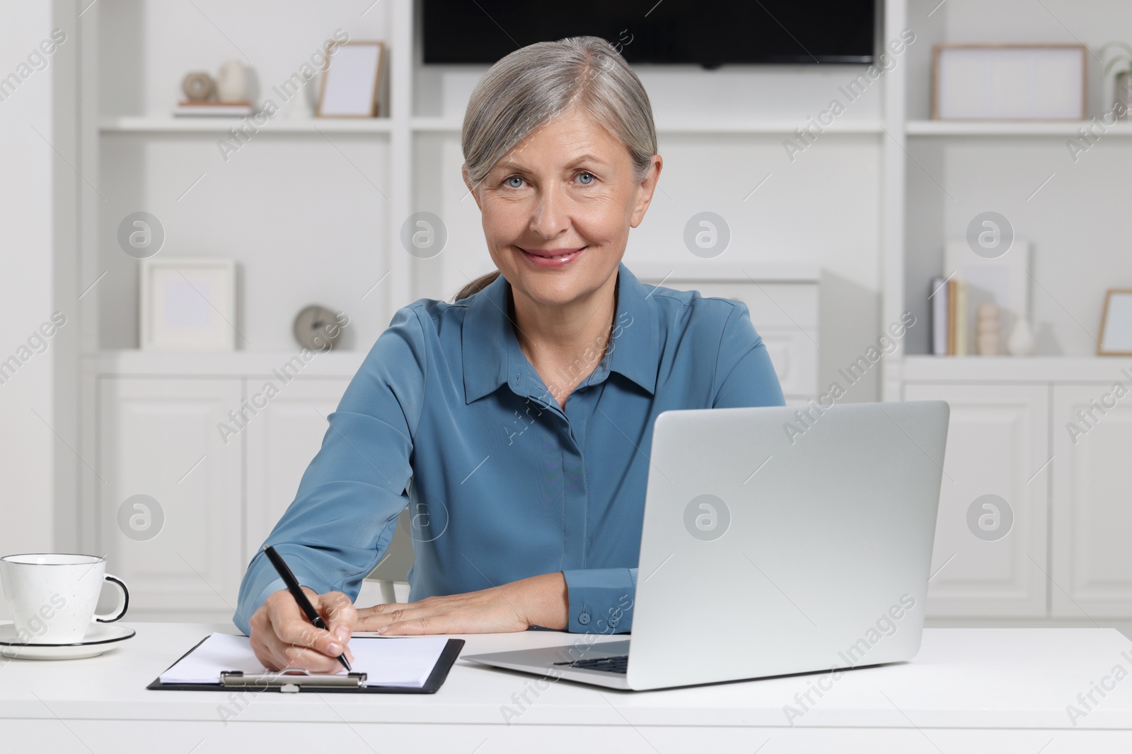 Photo of Beautiful senior woman taking notes near laptop at white table indoors