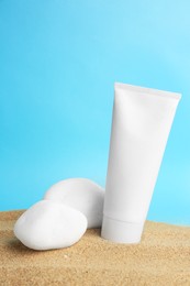 Photo of Tube of cream and stones on sand against light blue background. Cosmetic product