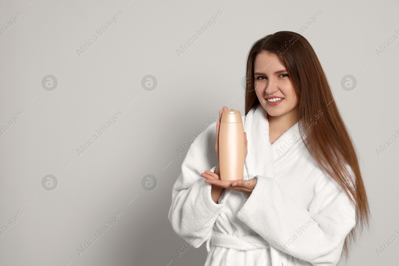 Photo of Beautiful young woman in bathrobe holding bottle of shampoo on light grey background. Space for text