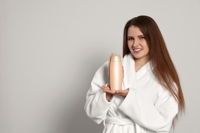 Beautiful young woman in bathrobe holding bottle of shampoo on light grey background. Space for text