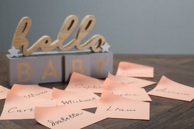 Photo of Cubes with phrase Hello Baby and paper stickers with different names on wooden table, closeup. Choosing baby's name