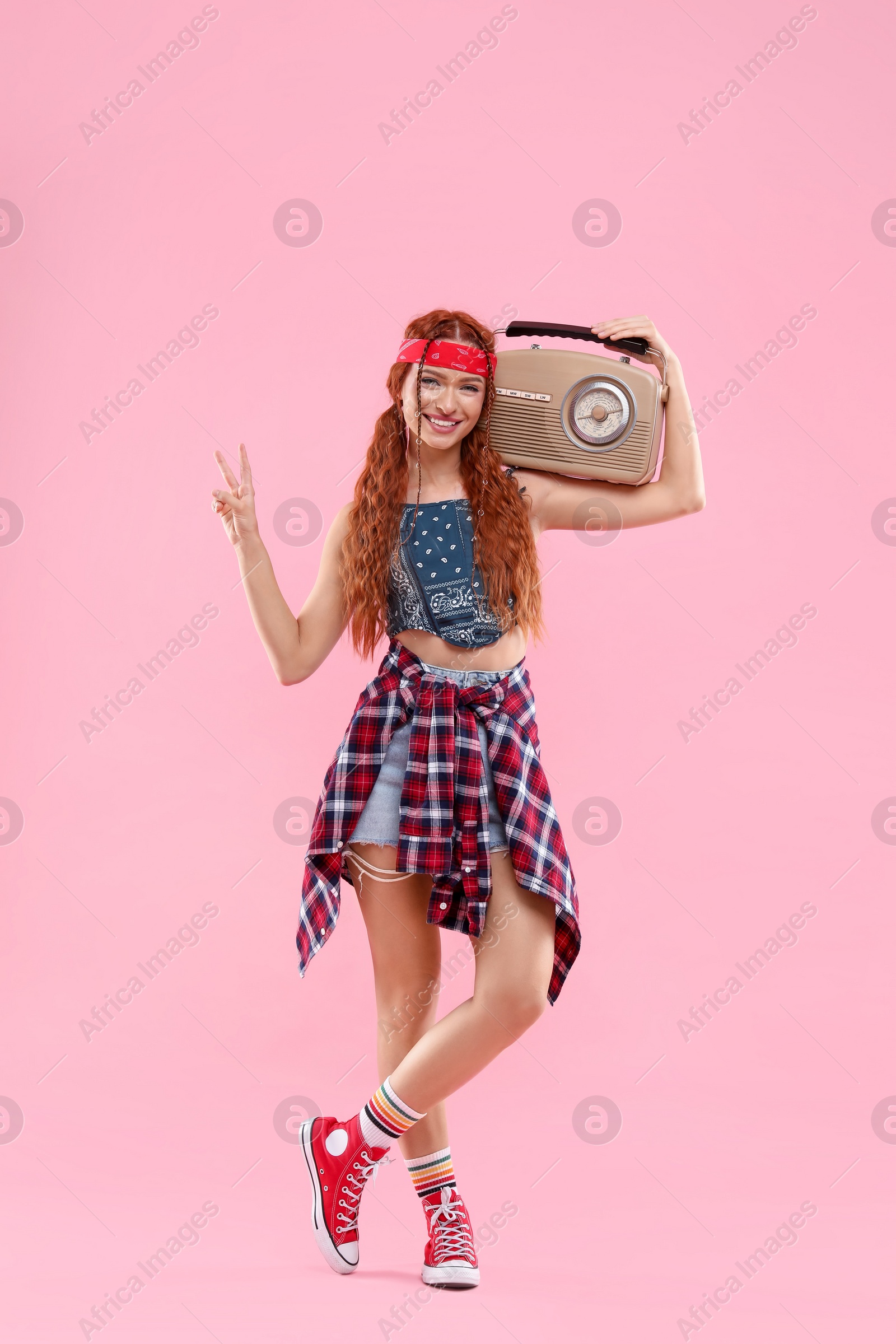 Photo of Stylish young hippie woman with radio showing V-sign on pink background