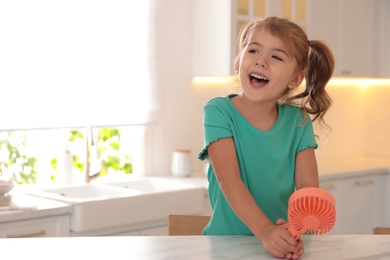 Photo of Little girl enjoying air flow from portable fan at table in kitchen. Summer heat