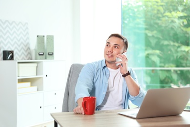 Photo of Young man talking on phone while working with laptop in home office