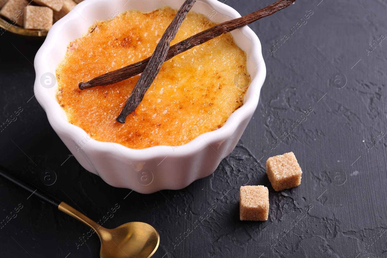 Photo of Delicious creme brulee in bowl, vanilla pods, sugar cubes and spoon on dark gray textured table, closeup