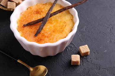 Photo of Delicious creme brulee in bowl, vanilla pods, sugar cubes and spoon on dark gray textured table, closeup