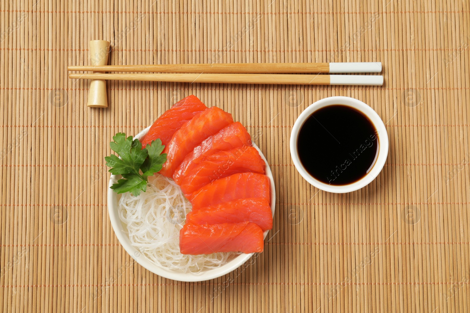 Photo of Delicious salmon sashimi served with funchosa, soy sauce and parsley on bamboo mat, flat lay