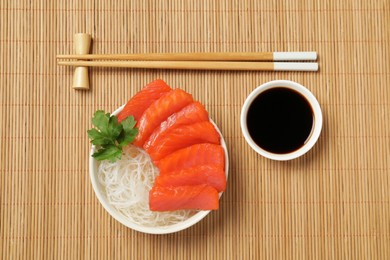 Photo of Delicious salmon sashimi served with funchosa, soy sauce and parsley on bamboo mat, flat lay