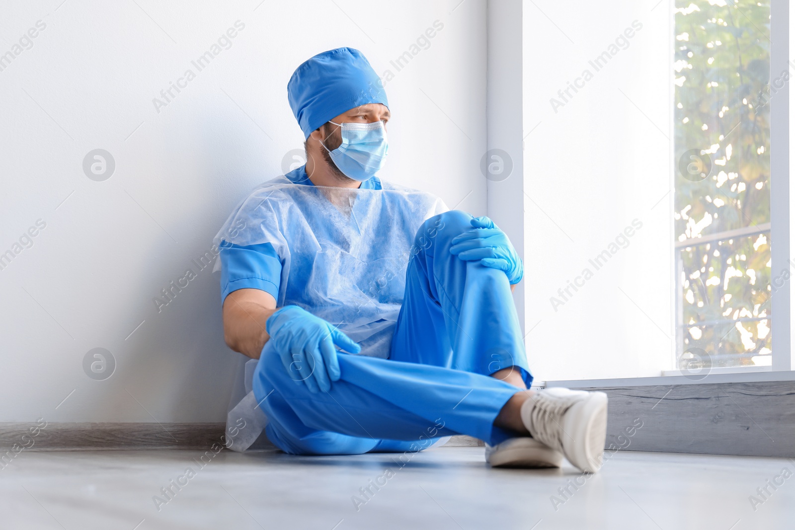 Photo of Exhausted doctor sitting on floor indoors. Stress of health care workers during COVID-19 pandemic