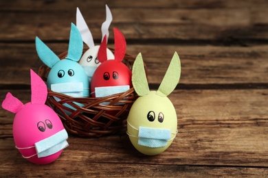 Photo of Painted eggs decorated with bunny ears and protective masks on wooden table, space for text. Easter holiday during COVID-19 quarantine