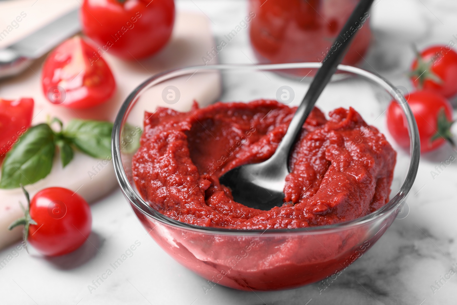 Photo of Glass bowl of tasty tomato paste with spoon and ingredients on white marble table, closeup