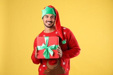 Photo of Happy man in Christmas sweater and elf hat holding gift box on yellow background