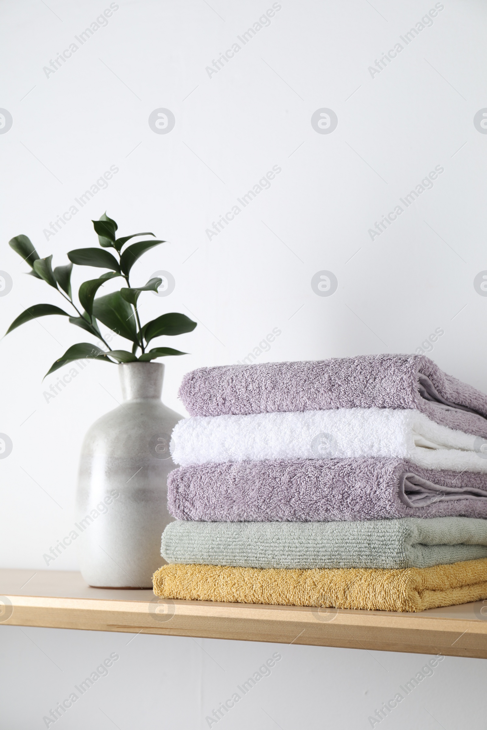 Photo of Stacked terry towels and green branches in vase on wooden shelf near white wall