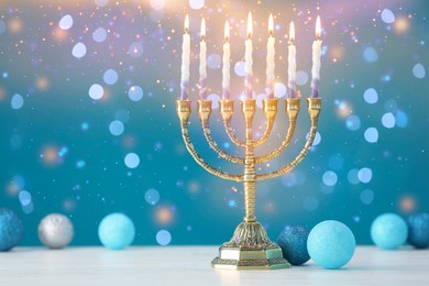 Image of Hanukkah celebration. Menorah with burning candles and baubles on white table against blue background with blurred lights, space for text