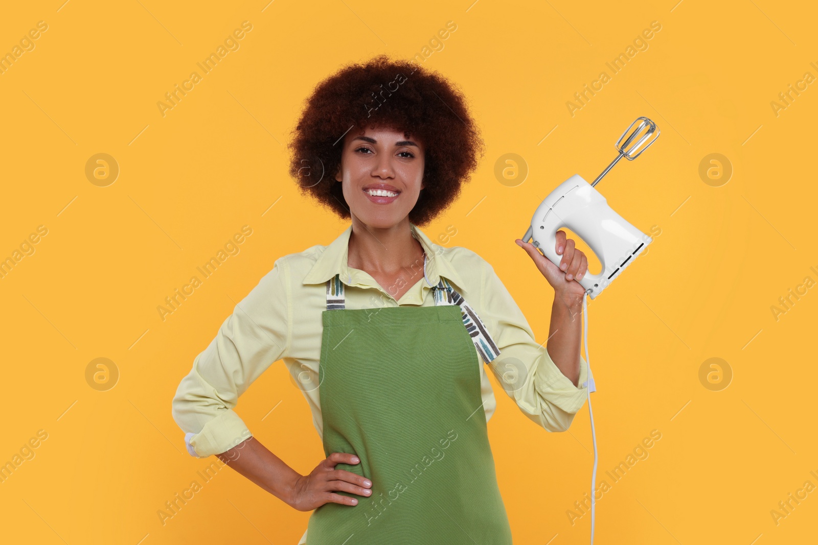 Photo of Happy young woman in apron holding mixer on orange background