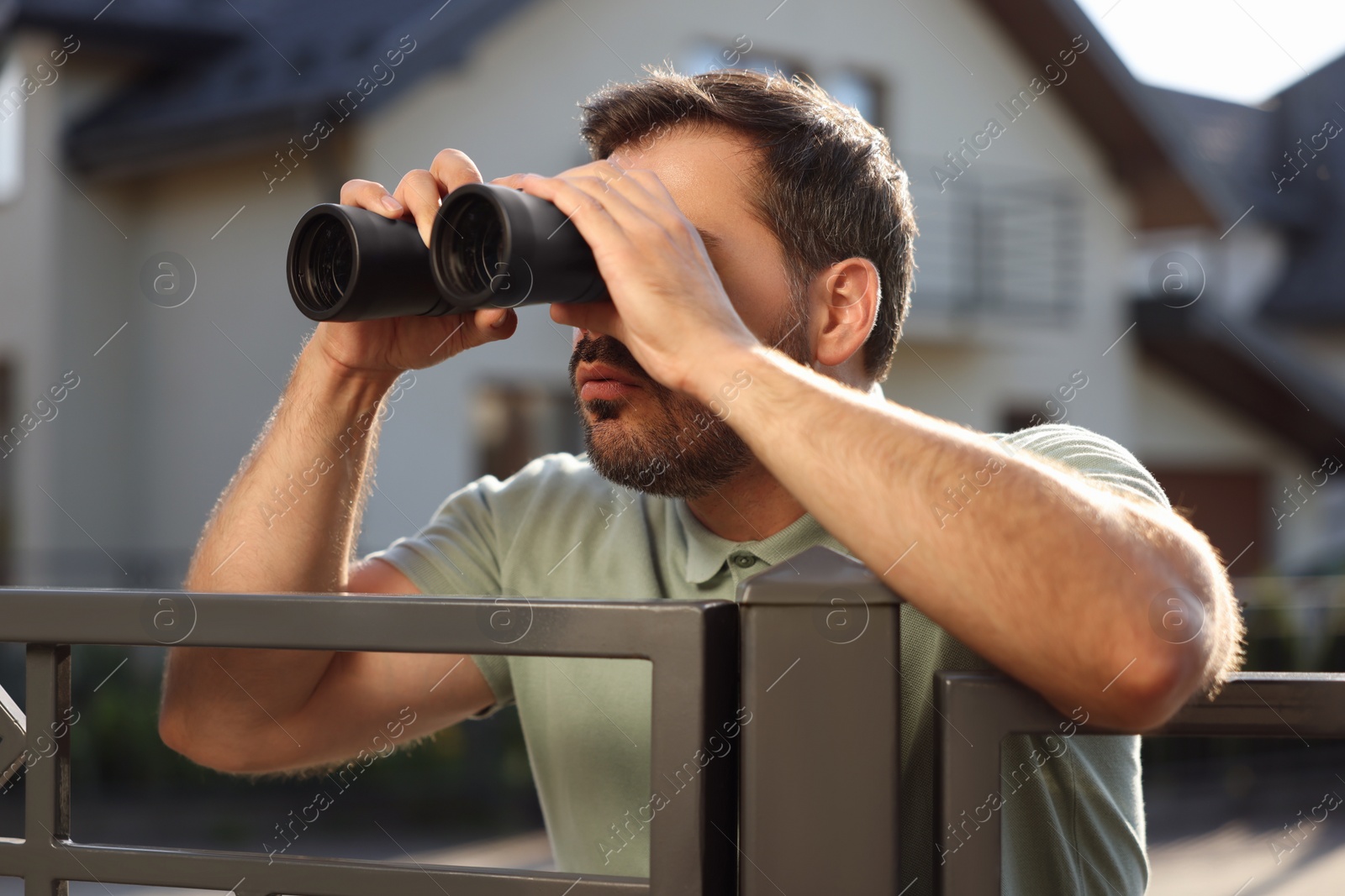 Photo of Concept of private life. Curious man with binoculars spying on neighbours over fence outdoors