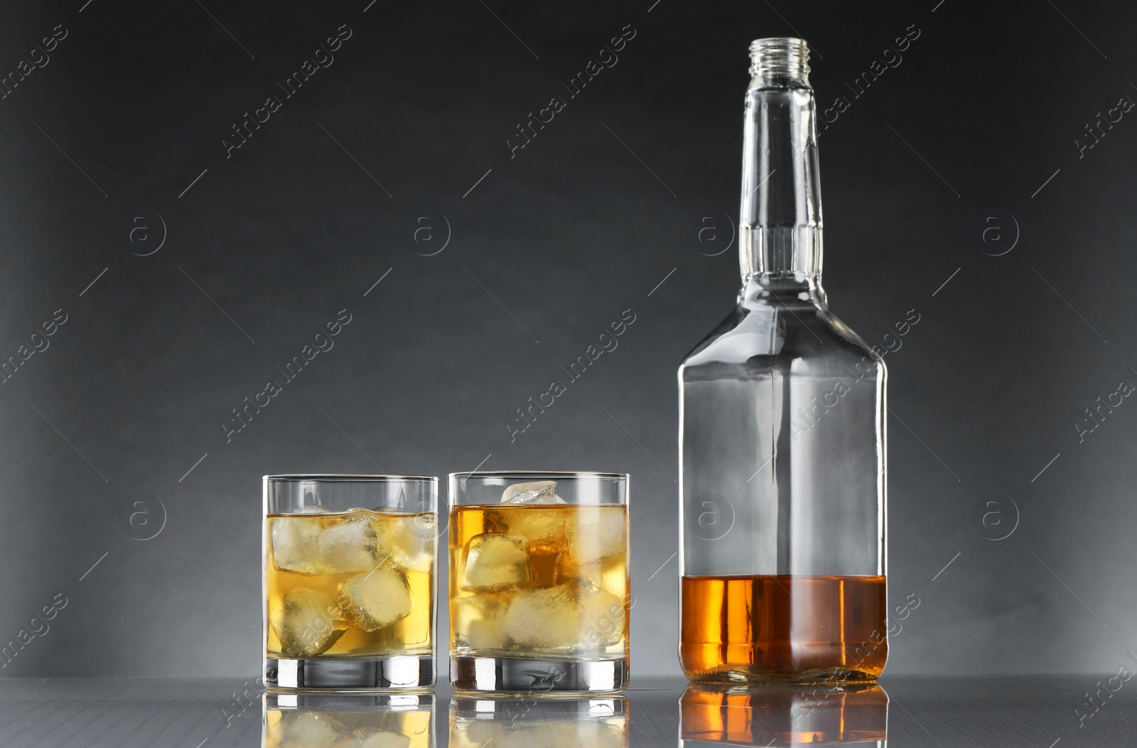 Photo of Tasty whiskey with ice in glasses and bottle on mirror table against dark background