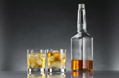 Tasty whiskey with ice in glasses and bottle on mirror table against dark background