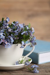 Photo of Beautiful forget-me-not flowers in cup, saucer and book on wooden table, closeup. Space for text