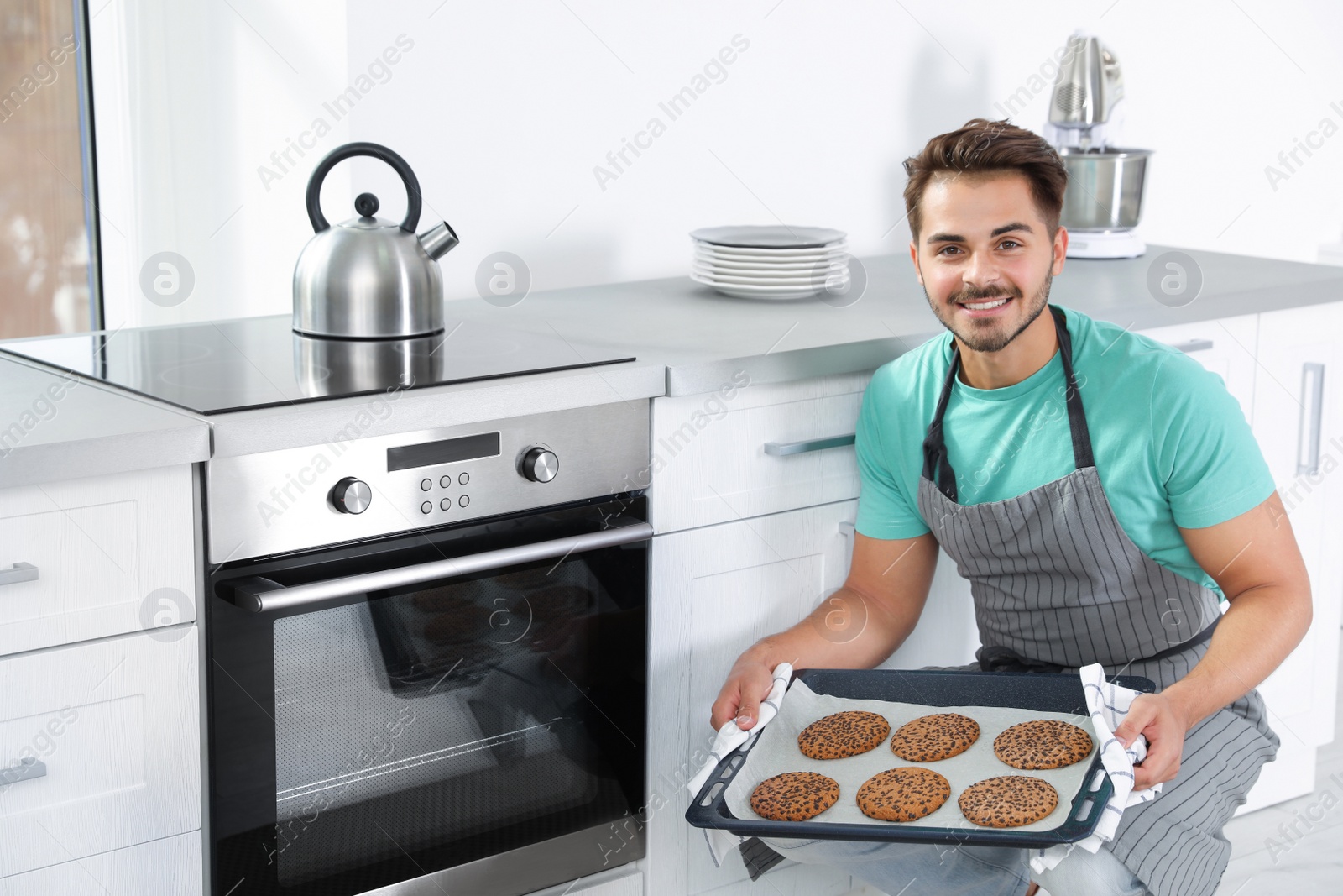 Photo of Young man holding baking sheet with cookies near oven in kitchen