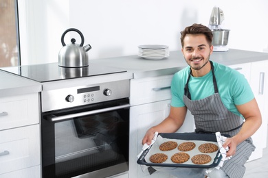 Young man holding baking sheet with cookies near oven in kitchen