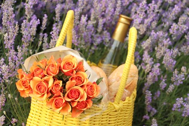 Yellow wicker bag with beautiful roses, bottle of wine and baguettes in lavender field, closeup