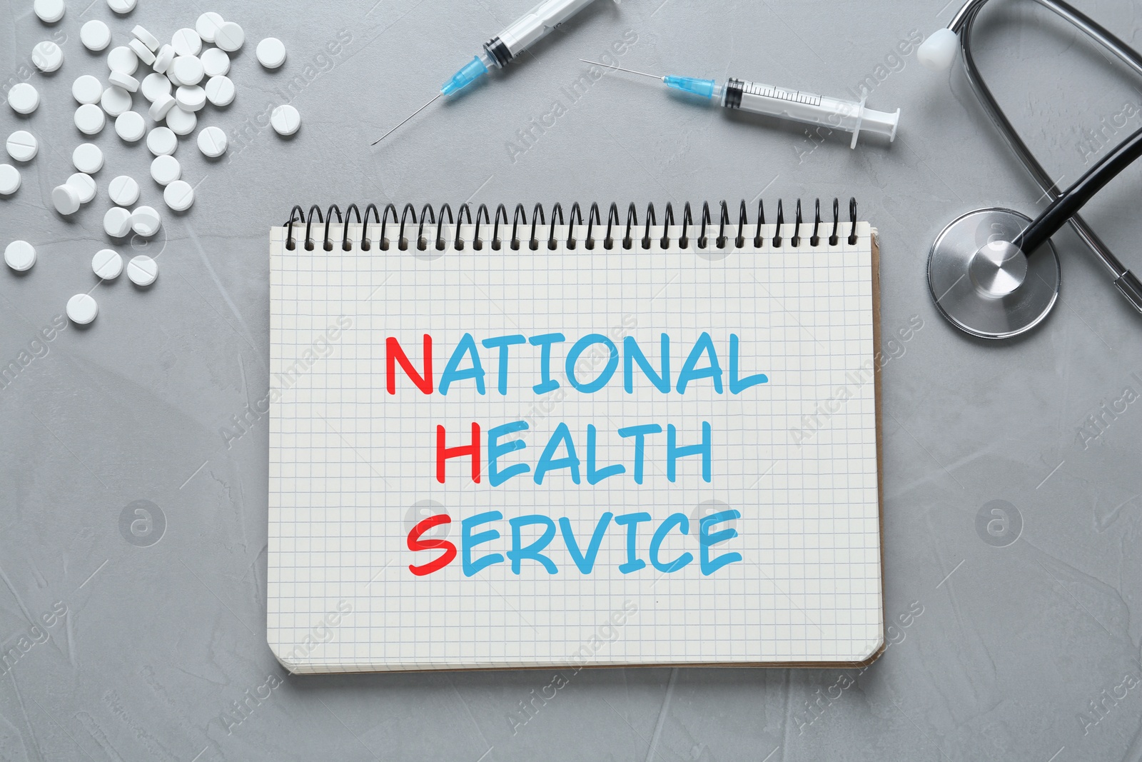 Image of National health service (NHS). Notebook with text, stethoscope, pills and syringes on grey background, flat lay