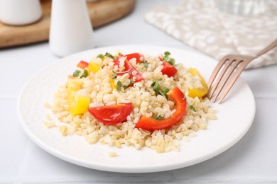 Plate of cooked bulgur with vegetables on white tiled table, closeup