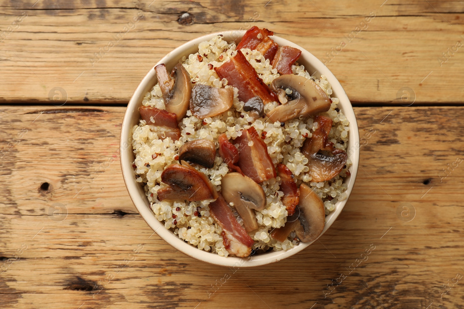 Photo of Tasty quinoa porridge with fried bacon and mushrooms in bowl on wooden table, top view