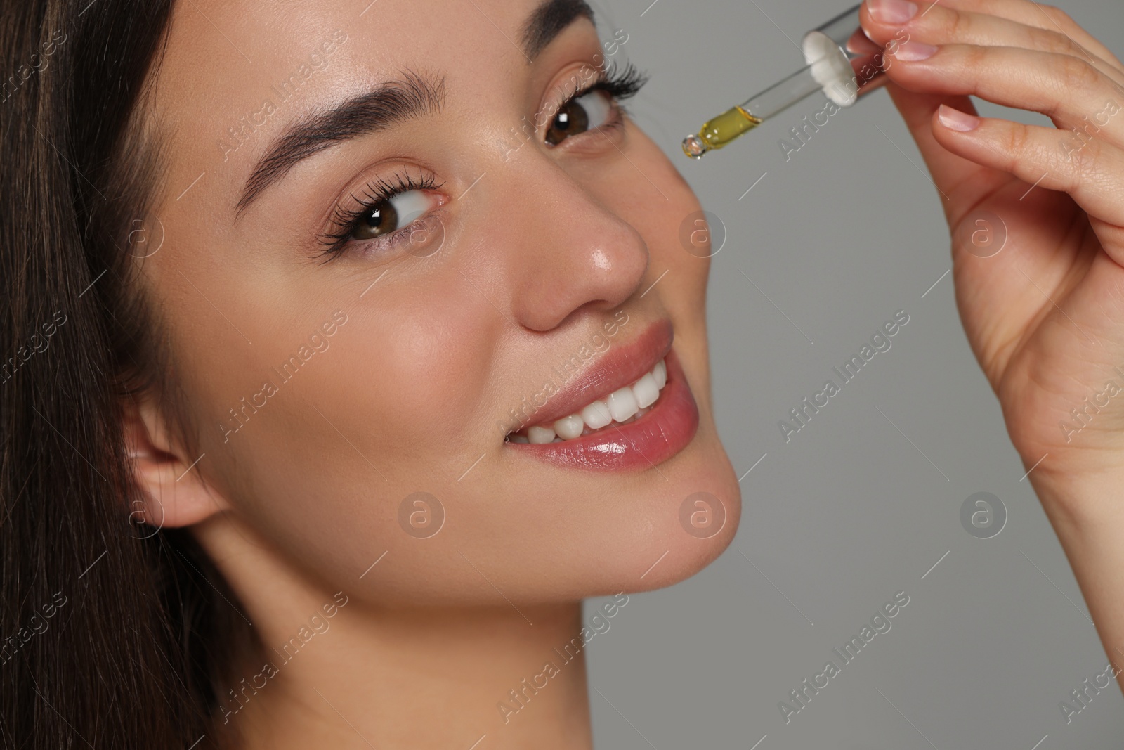 Photo of Beautiful young woman applying essential oil onto face on grey background, closeup